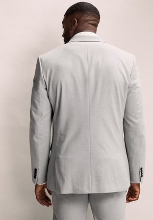 Homme de dos portant une veste de costume gris clair avec un pantalon assorti sur un fond beige uni.