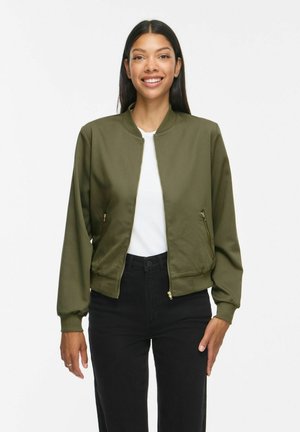 Femme souriante aux longs cheveux foncés portant une veste bomber vert olive, une chemise blanche et un pantalon noir taille haute, debout devant un fond blanc.