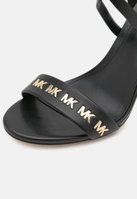 Czarna skórzana sandały z złotymi ćwiekami w kształcie logo MK na przednim pasku oraz cienkim paskiem na kostce, przedstawione na białym tle.