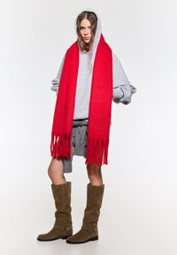 Rode franjesjaal, grijze oversized hoodie, gestructureerde grijze shorts en bruine suede kniehoge laarzen met banddetails. Effen witte achtergrond.