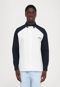 Uomo che indossa una camicia bianca con bottoni e maniche e colletto neri, e pantaloni a righe bianche e azzurre, in piedi davanti a uno sfondo neutro.