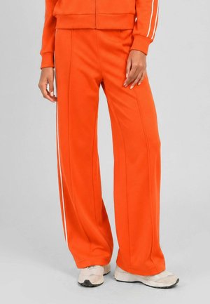 Personne portant un pantalon large orange vif avec des bandes blanches sur les côtés et des baskets blanches, debout devant un fond gris uni.