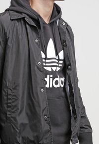 Svart, lätt jacka med tryckknappar över en grå hoodie med en vit adidas-logotyp och det ikoniska tre-stripes-designet.