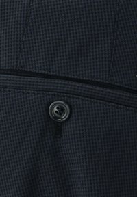 Dunkel gemustertes Gewebe mit einem dezenten Hahnentrittmuster, einem einzelnen schwarzen Knopf und einem Reißverschlussdetail auf der linken Seite.