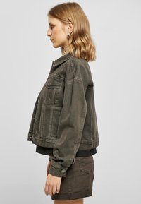 Olive green denim jacket with a classic collar, front pockets, and silver-tone buttons, paired with a black shirt and brown shorts.