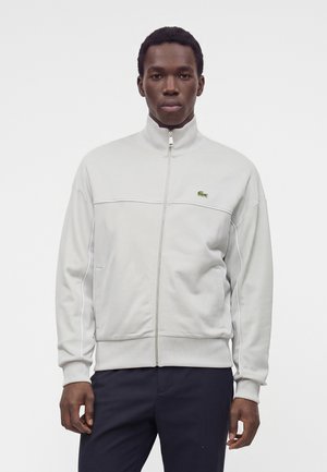 Homme portant une veste de survêtement gris clair zippée avec un logo crocodile vert et un pantalon foncé, debout devant un fond blanc uni.