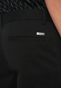 Podrobni posnetek zadnjega žepa črnih hlač z metalno oznako Armani Exchange in črno vzorčasto srajco, vstavljeno notri.