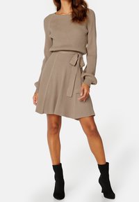 Frau, die ein beige geripptes Strickkleid mit langen Ärmeln, einem Taillengurt und schwarzen Knöchelstiefeln vor einem weißen Hintergrund trägt.