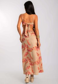 Mujer con cabello largo y oscuro que lleva un vestido maxi beige con patrones de hojas en rosa y naranja, escote en la espalda, lazo atado y sandalias de cuña.