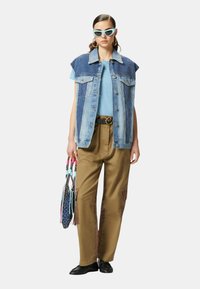 Gilet di denim con spalline, t-shirt blu, pantaloni marroni con tasche laterali e scarpe nere. Gli accessori includono borse colorate e occhiali da sole.