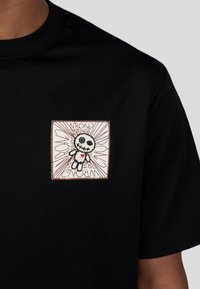 Czarna bawełniana koszulka z kwadratową łatą przedstawiającą postać w stylu kreskówkowym, otoczoną napisem „WRONG FRIENDS” w formie wybuchowego designu.