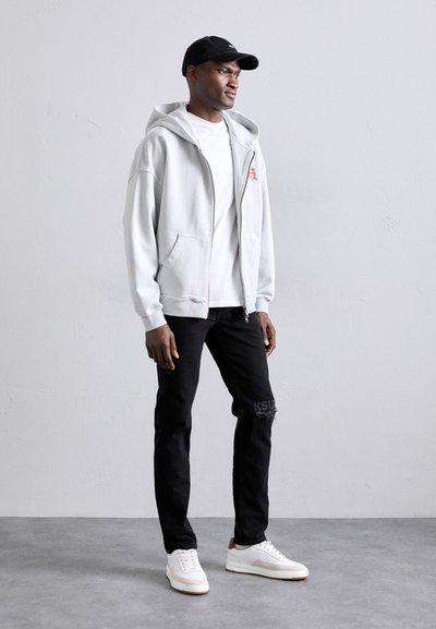 Ljusgrå zip-tröja med ficka i känguru, bärs över en vit skjorta, ihop med svarta jeans och vita sneakers. Svart keps som bärs på sned.