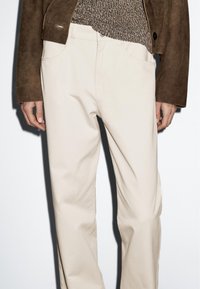 Béžové bavlněné kalhoty s rovným střihem, opatřené předními kapsami a čistým designem, v kombinaci s texturovaným hnědým svetrem.