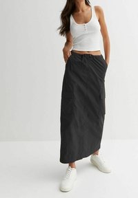 Vrouw in een witte mouwloze ribbeltop, lange zwarte rok met zakken en witte sneakers, staand met handen in de zakken.