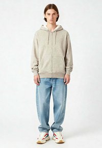 Felpa zip grigia chiara con cappuccio foderato in felpa, dotata di cordini e tasche frontali, abbinata a jeans blu larghi e sneakers color crema.