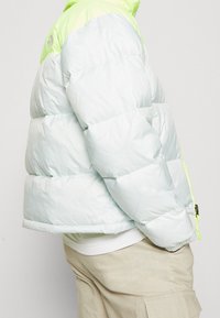 Jasnoniebieska kurtka puchowa z zielonym kapturem, posiadająca pikowany wzór i widoczne elementy zamka błyskawicznego. Miękka tekstura, lekkie materiały.