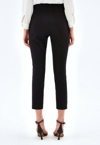 Vrouw die staat met zwarte, strakke, enkellange broek, witte blouse met lange mouwen en zwarte, hoge hakken, gezien vanuit de rug.
