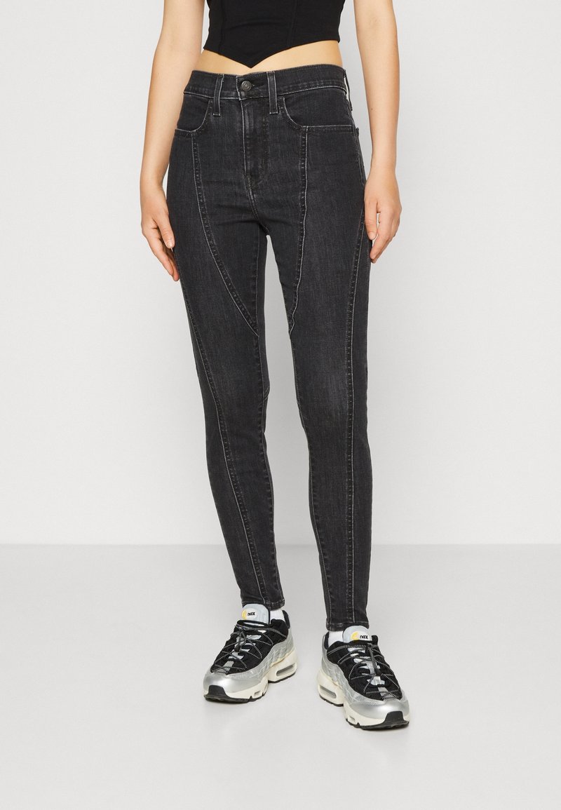 Personne portant un jean skinny noir taille haute avec des coutures contrastantes et des baskets noires et argentées, debout devant un fond uni.