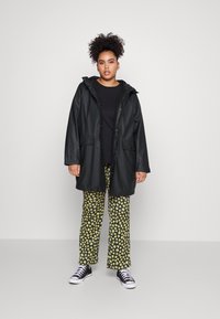 Schwarze wasserdichte Jacke mit Kapuze, kombiniert mit schwarzem Oberteil und Hose mit Blumendruck. Das Model trägt schwarze Sneakers. Einfarbiger Hintergrund.