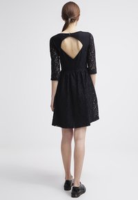 Schwarzes Spitzenkleid mit Dreiviertelärmel, tailliertem Oberteil, ausgestelltem Rock und einem dreieckigen Ausschnitt am Rücken. Ein Paar schwarze Schuhe ist sichtbar.
