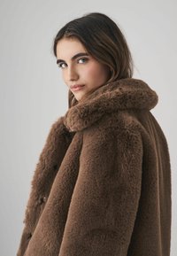Brun faux fur frakke med en bred krave, der har en blød tekstur og en afslappet pasform. Frakken har subtile knapdetaljer og et enkelt design.