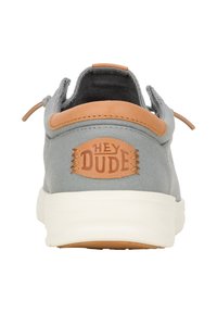 Zapatilla de tela gris con un parche de cuero marrón que lleva la marca "HEY DUDE", suela blanca y forro textil. Diseño minimalista con detalle de cordones.
