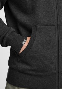 Sudadera gris con cremallera, hecha de un tejido suave, que cuenta con un bolsillo frontal, puños acanalados y una pequeña etiqueta de logo cerca de la manga.