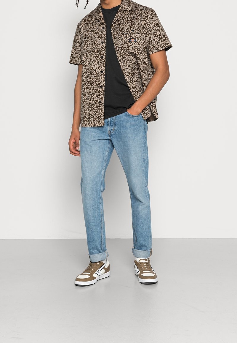 Camisa de leopardo de manga corta, camiseta negra, jeans de color azul claro y zapatillas marrones/blancas llevadas por una figura de pie contra un fondo blanco.