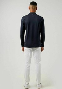 Marineblaues langärmliges Poloshirt mit hohem Kragen, kombiniert mit weißen Slim-Fit-Hosen und weißen Sneakern. Glatter Stoff, minimalistisches Design.
