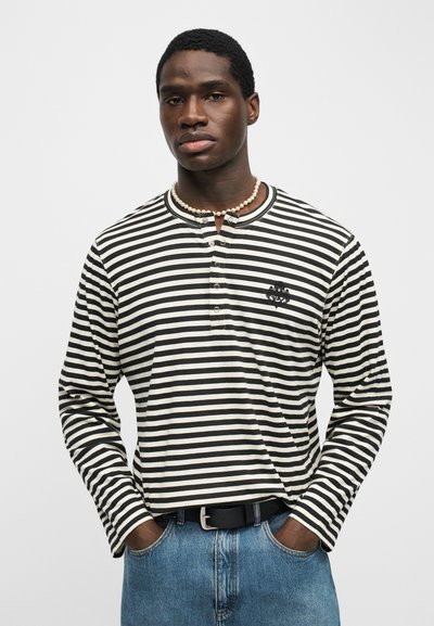 Νεαρός άνδρας που φοράει μακρυμάνικη ριγέ μαύρο-λευκό πουκάμισο, μαργαριταρένιο κολιέ, μπλε τζιν και έχει τα χέρια στις τσέπες, κοιτάζοντας μπροστά.