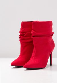 Botines de ante rojos con un diseño holgado, punteras puntiagudas y tacones de aguja. Textura suave sin patrones ni elementos metálicos visibles.