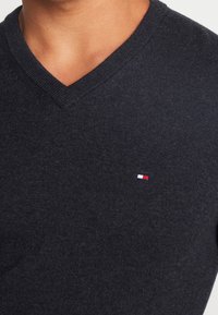 Czarny szary sweter w serek wykonany z miękkiego materiału dzianinowego, z małym, trójkolorowym logo na klatce piersiowej. Minimalistyczny design, gładka faktura.