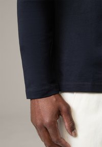 Navy long-sleeve shirt with a smooth texture, fitted design, and stitched cuffs. Hand resting on white fabric below the shirt.