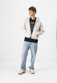 Haut de survêtement zippé gris clair avec une grande poche avant, porté sur un t-shirt graphique noir, associé à des jeans bleu clair et des baskets blanches.