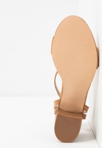 Sandale en daim beige avec une fine bride à la cheville, un bout ouvert et une semelle en caoutchouc texturée avec une finition lisse. Le talon mesure environ 3 pouces.