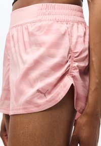 Κοντινό πλάνο ενός ατόμου που φοράει ανοιχτό ροζ αθλητικά σορτς Puma με διακριτικά αφηρημένα σχέδια και πλαϊνές σχισμές, που δείχνει το πάνω μέρος του μηρού και το χέρι του.