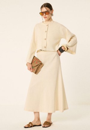 Femme portant un haut crème à manches longues boutonné et une longue jupe assortie, tenant une pochette tissée, avec de grandes lunettes de soleil ambrées oversize et des sandales.