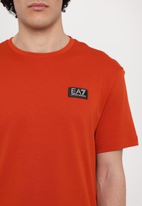 Red cotton t-shirt with crew neckline, short sleeves, and a black fabric patch displaying “EA7 Emporio Armani” on the chest.