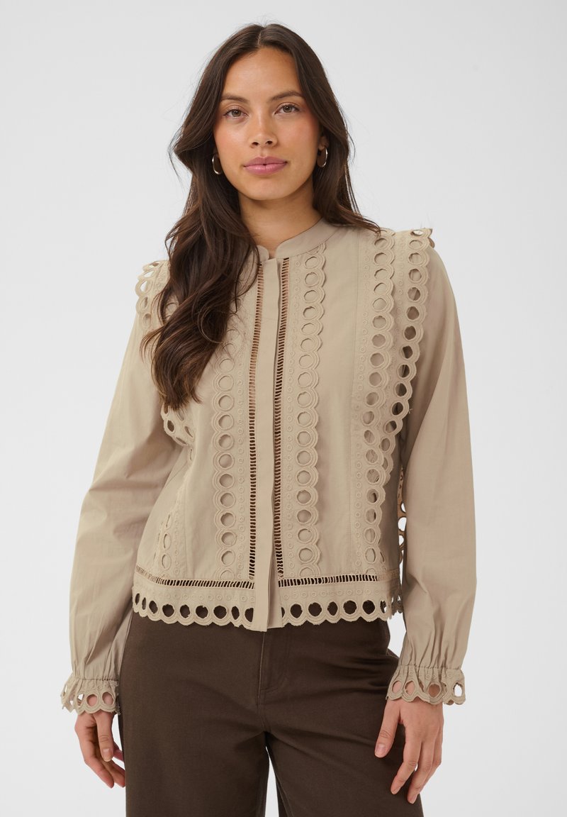 Femme portant un chemisier beige à manches longues avec des détails en dentelle circulaire et un pantalon marron foncé, debout devant un fond clair et uni.