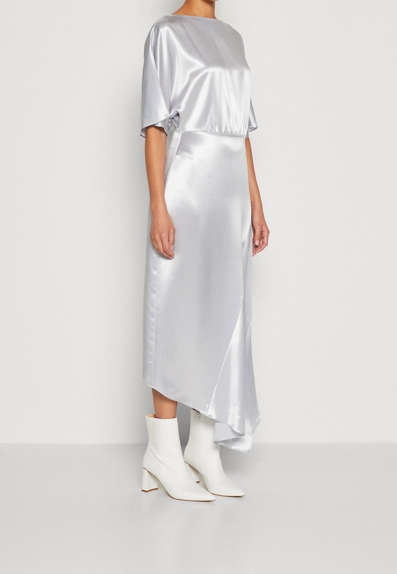 Robe midi en satin argenté avec un décolleté bateau et des manches courtes, présentant un ourlet asymétrique, associée à des bottes à talons blanches.