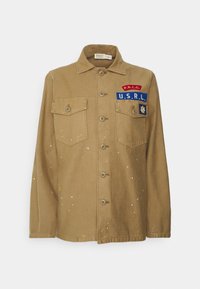 Chaqueta de lona caqui con dos bolsillos en el pecho, cierre de botones y diversas manchas de pintura colorida. Incluye parches bordados.
