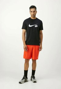 Hombre con camiseta negra de Nike, pantalones cortos naranja brillante, calcetines negros y zapatillas grises, de pie contra un fondo blanco sencillo.