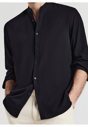 Homme portant une chemise noire à col mandarin boutonnée avec les manches roulées et un pantalon beige, mains dans les poches, sur un fond clair.