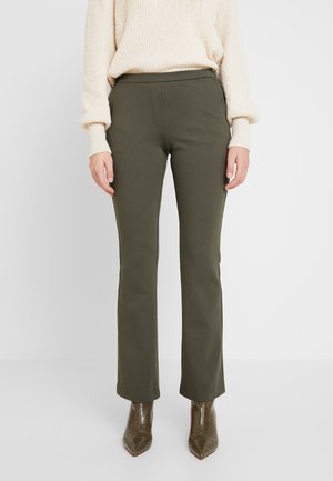 Femme portant un pantalon droit vert olive, un pull en maille beige et des bottines vert olive à bout pointu, debout sur fond blanc.