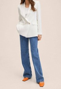 Kobieta ubrana w białą dwurzędową marynarkę, jasny top, niebieskie szerokie spodnie i pomarańczowe buty na szpilce, stojąca na tle jednolitego tła.