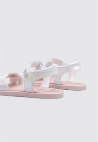 Sandalias blancas con un acabado brillante, suela rosa, correa ajustable y acentos texturizados. Diseño abierto con plantilla moldeada para mayor comodidad.