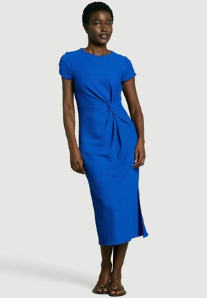 Frau trägt ein hellblaues Midi-Kleid mit kurzen Ärmeln, seitlichem Knoten und Schlitz, steht vor einem einfarbigen grauen Hintergrund.