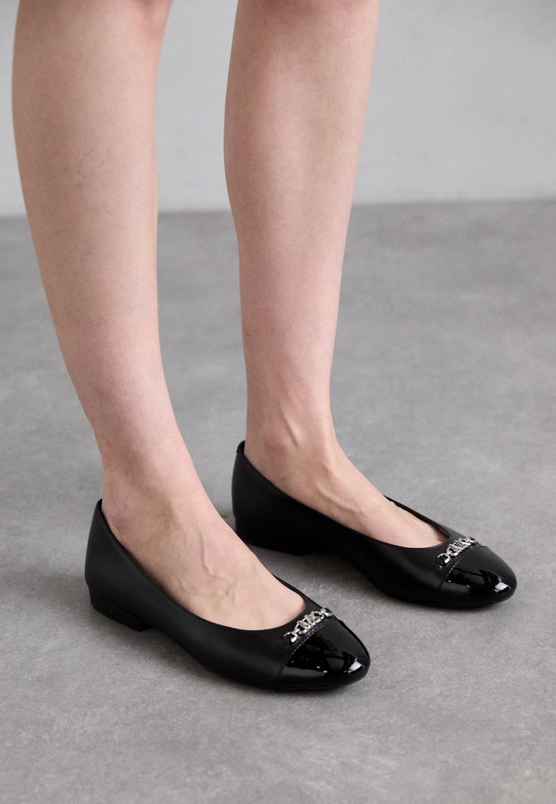 Chaussures plates noires avec bouts en cuir verni et détail chaîne argentée, portées pieds nus, debout sur un sol gris.