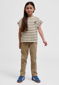 Stribet beige top med volanger på ærmerne, parret med khaki bukser og blå sneakers med hvide accenter; broderet logo på skjorten og bukserne.