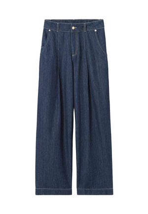 Femme debout portant une veste marron à carreaux, un pantalon large en jean bleu, des chaussures blanches et une écharpe bleue avec une bordure rouge autour du cou.